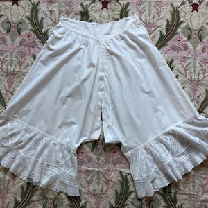 May include: White cotton bloomers featuring a wide, gathered waistband and a V-shaped front seam. The legs are loose and wide, ending in flared, ruffled hems decorated with intricate white lace embroidery and detailed eyelet patterns.