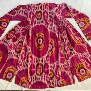 May include: A long, flowing garment in shades of fuchsia, orange, and cream. The fabric features a repeating pattern of circular and oval shapes. The sleeves are long and the garment is open in the front, creating a dramatic silhouette. The textile has a slightly textured appearance.