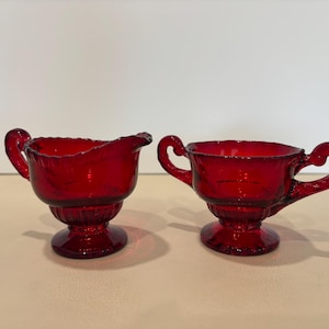 May include: A pair of ruby red glass vessels, including a creamer with a pouring spout and a sugar bowl with two side handles. Both pieces feature a pedestal base, a scalloped rim, and a ribbed texture on the lower body, crafted from translucent red glass.