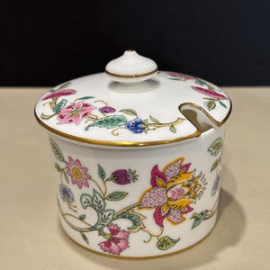 Op de afbeelding: Een witte porseleinen jam-pot met bijpassend deksel voorzien van een kleurrijk bloemmotief in roze, geel, blauw en groen. De pot heeft een goudkleurige rand en een kleine inkeping in het deksel voor een lepel.
