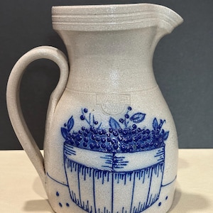 May include: A light grey stoneware pitcher featuring a blue hand-painted design of a basket filled with blueberries. The pitcher has a curved handle, a pouring spout, and a stamped logo that reads Smiths Falls Stoneware 2003 on the front.