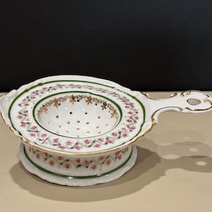 May include: A white porcelain tea strainer with a handle, featuring a delicate pink rose and green ribbon pattern around the rim and base. The bowl of the strainer has small drainage holes and gold-painted accents along the edges and rim.