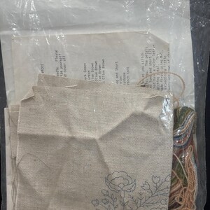 Op de afbeelding: Een borduurpakket in een doorzichtige plastic zak met een stuk beige stof met een blauwe bloemenomtrek, een gedrukt instructieblad met de tekst #A302 en een bundel met diverse kleuren borduurgaren.