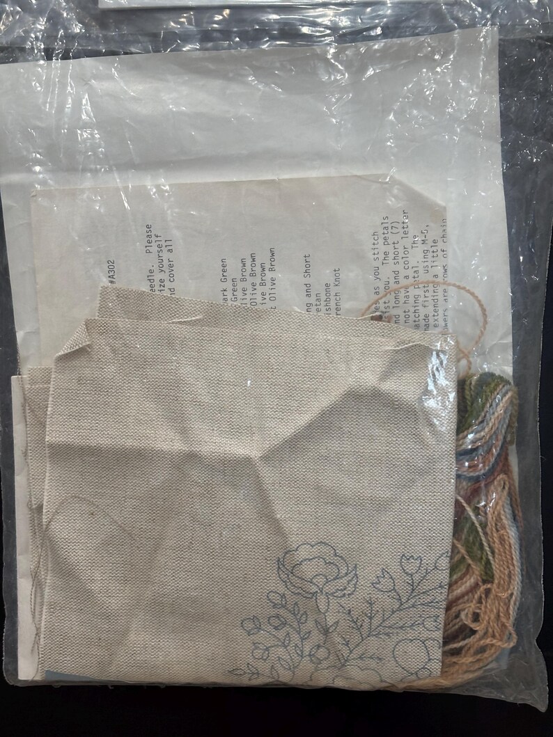 Op de afbeelding: Een borduurpakket in een doorzichtige plastic zak met een stuk beige stof met een blauwe bloemenomtrek, een gedrukt instructieblad met de tekst #A302 en een bundel met diverse kleuren borduurgaren.
