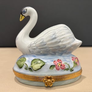 Op de afbeelding: Een porseleinen snuisterijdoosje met een witte zwaanfiguur op het deksel. De basis is beschilderd met lichtblauw water, groene bladeren en roze bloemen. Een goudkleurig metalen scharnier met een bloemvormige sluiting houdt het doosje dicht.