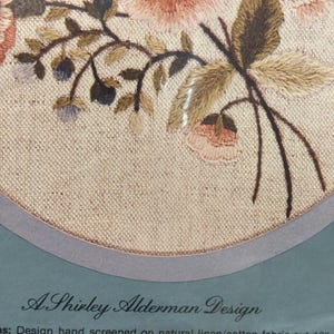 Op de afbeelding: Een borduurpakket met een bloemmotief met roze en perzikkleurige bloemen en groene bladeren op natuurlijk linnen. De verpakking bevat de tekst A Shirley Alderman Design en vermeldt dat de stof is gesneden op 30,48 cm bij 30,48 cm.