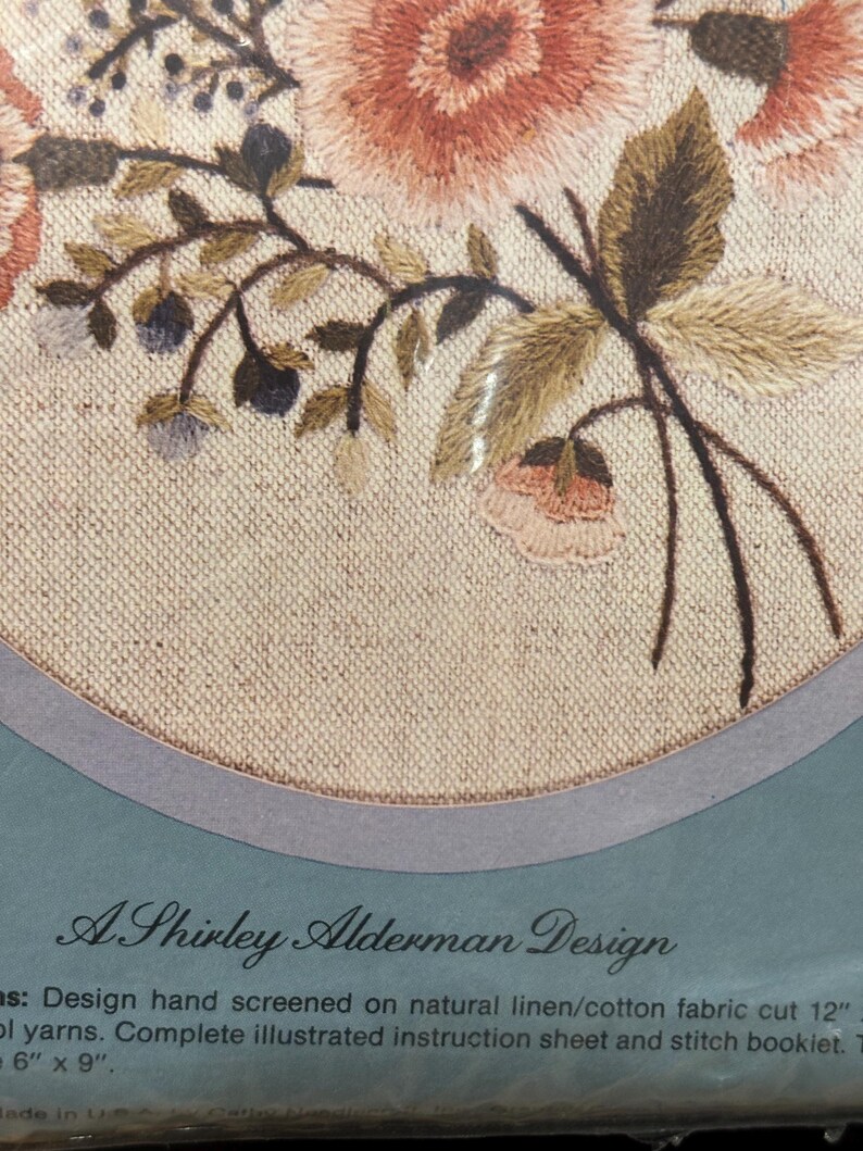Op de afbeelding: Een borduurpakket met een bloemmotief met roze en perzikkleurige bloemen en groene bladeren op natuurlijk linnen. De verpakking bevat de tekst A Shirley Alderman Design en vermeldt dat de stof is gesneden op 30,48 cm bij 30,48 cm.