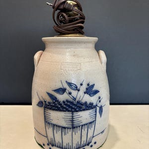 May include: A beige stoneware lamp base with two side handles, featuring a blue hand-painted design of a basket filled with berries and leaves. The text SALMON FALLS STONEWARE 1993 is stamped on the front. A brown electrical cord is coiled on top.