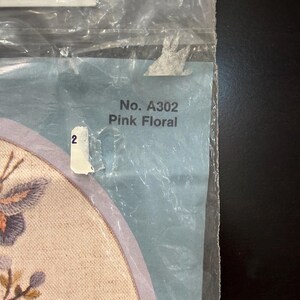 Op de afbeelding: Een doorzichtige plastic verpakking met een knutselartikel op een lichtgekleurde stoffen ondergrond. De verpakking bevat de tekst No. A302 Pink Floral en een klein wit label met het cijfer 2. Een deel van een geborduurd bloemmotief is zichtbaar.
