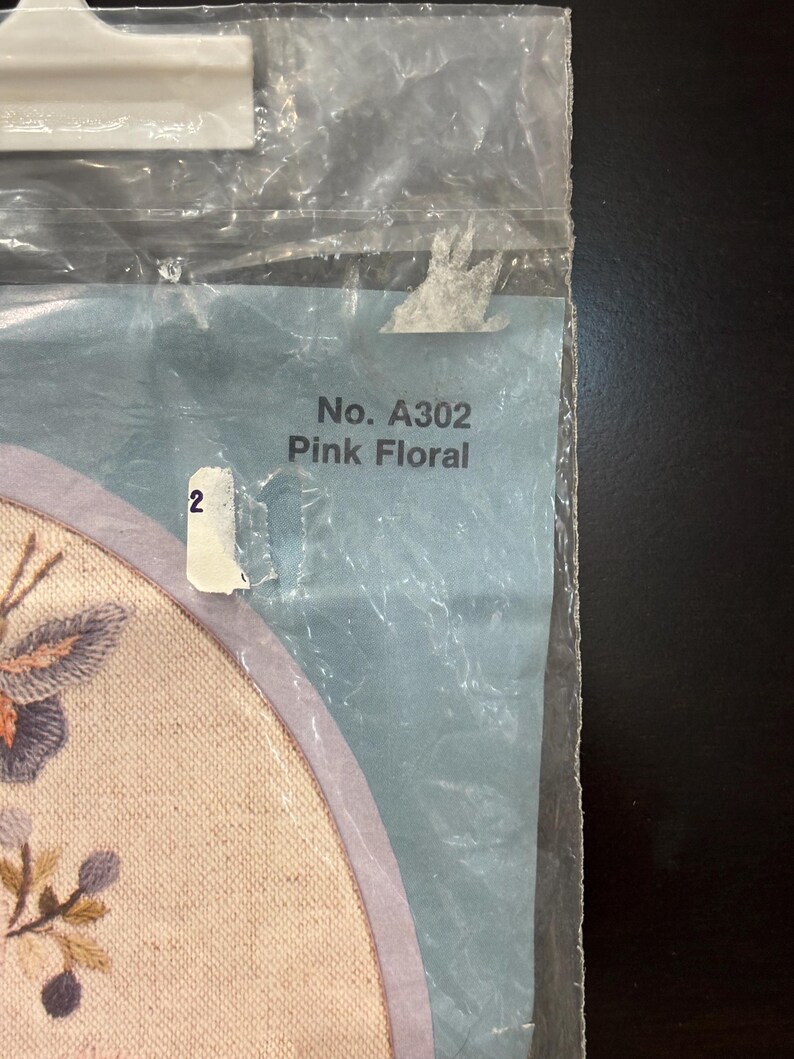 Op de afbeelding: Een doorzichtige plastic verpakking met een knutselartikel op een lichtgekleurde stoffen ondergrond. De verpakking bevat de tekst No. A302 Pink Floral en een klein wit label met het cijfer 2. Een deel van een geborduurd bloemmotief is zichtbaar.