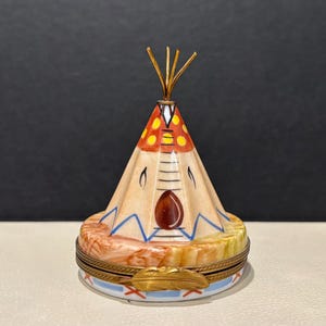 Op de afbeelding: Een porseleinen snuisterijendoosje in de vorm van een tipi met een kegelvormig dak met oranje en gele cirkelvormige patronen. De basis is rond met een gemarmerde textuur en een goudkleurige metalen sluiting in de vorm van een veer.