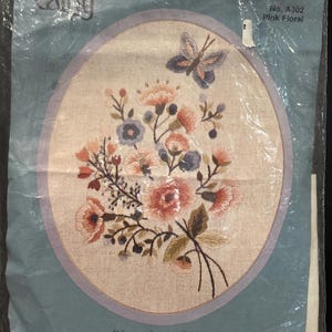 May include: A needlepoint kit titled Pink Floral, No. A302, featuring a Shirley Alderman design. The kit includes a natural linen/cotton fabric with a hand-screened floral and butterfly pattern, crewel wool yarns, two needles, and instructions.