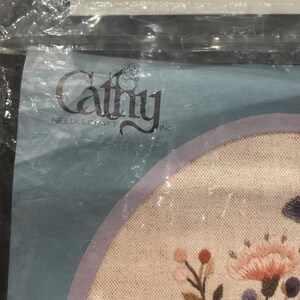 Op de afbeelding: Een Cathy Needlecraft Inc. borduurpakket met een rond stuk stof met geborduurde bloemmotieven in roze, blauwe en groene tinten, verpakt in een doorzichtige plastic zak met een witte plastic hanger aan de bovenkant.