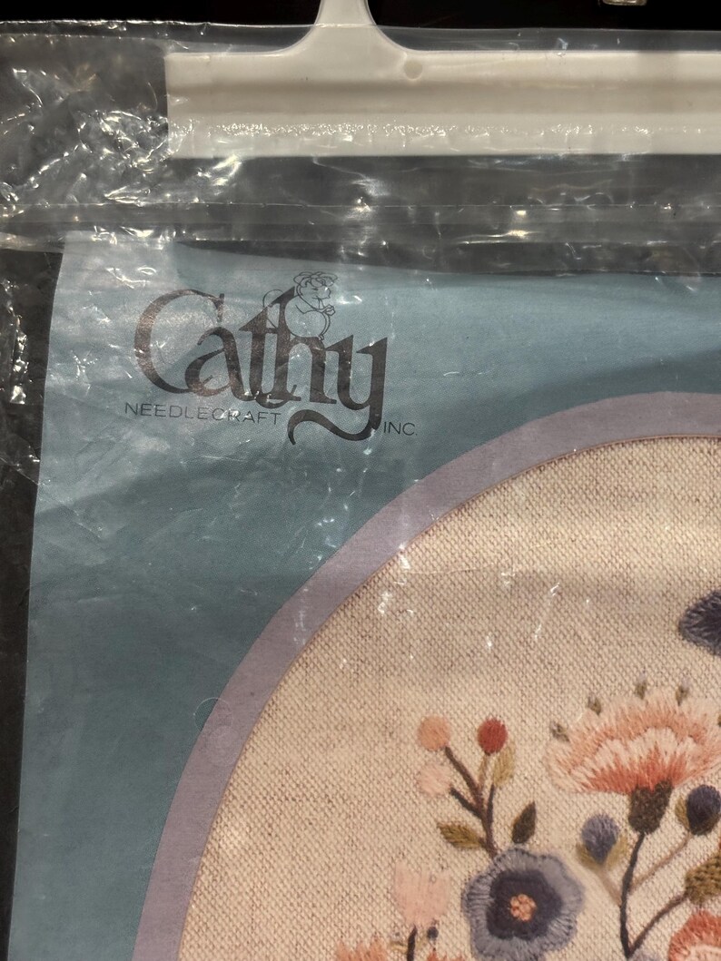 Op de afbeelding: Een Cathy Needlecraft Inc. borduurpakket met een rond stuk stof met geborduurde bloemmotieven in roze, blauwe en groene tinten, verpakt in een doorzichtige plastic zak met een witte plastic hanger aan de bovenkant.
