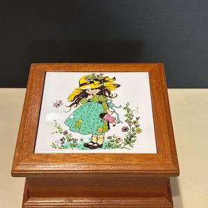 Op de afbeelding: Een vierkante houten doos met een keramische tegel in de deksel. De tegel toont een kleurrijke illustratie van een persoon met een gele hoed en een groene jurk, die een kleine pop vasthoudt, omringd door bloemen, een lieveheersbeestje en een dier.