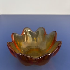 Pode incluir: Uma pequena taça de vidro translúcido cor de âmbar com uma borda recortada em forma de pétalas. A taça apresenta um interior liso e curvo, com um tom laranja-acastanhado profundo que transita para um tom dourado mais claro no centro.