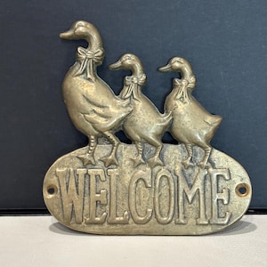 May include: A brass wall plaque featuring a large duck followed by two smaller ducklings, each wearing a bow around its neck. The word WELCOME is embossed in capital letters on the oval base, which includes two small holes for mounting purposes.