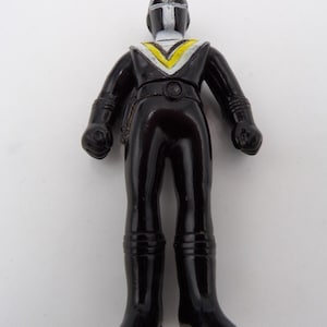 May include: A black plastic action figure with a smooth finish, featuring a white and yellow V-shaped chest emblem, a white vertical stripe on the helmet, and molded details on the arms and legs, standing in a neutral pose with clenched fists.