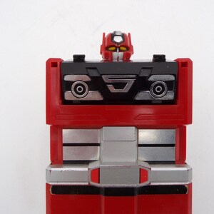 May include: A red and black plastic robot action figure with a blocky, rectangular torso. The head is red with yellow eyes and a silver crest. The chest features a black panel with silver circular details and a central silver emblem. The lower section has horizontal silver and red stripes.