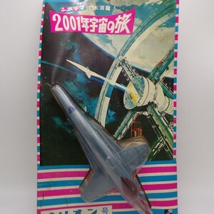 Pode incluir: Um modelo de nave espacial de plástico cinzento na embalagem original. O cartão apresenta uma ilustração de uma estação espacial e o texto japonês 2001年宇宙の旅 e オリオン号. A embalagem tem riscas horizontais cor-de-rosa e azuis na parte superior e inferior.