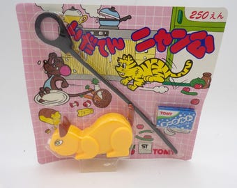 May include: A yellow plastic cat toy packaged on a pink grid-patterned cardboard backing. The packaging features a cartoon cat, a mouse, and kitchen imagery with Japanese text. A black plastic tool is included, along with a small blue Tomy gum box.