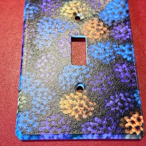 May include: A rectangular light switch wall plate featuring a dense, textured pattern of small flowers in shades of blue, purple, and orange against a black background. The plate has a central rectangular cutout and two circular screw holes.