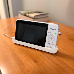 May include: A white VTech baby monitor with a rectangular black screen and a control panel on the right side featuring navigation buttons, a power button, a talk button, and volume controls. A green indicator light is illuminated above the buttons.