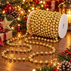 May include: A spool of metallic gold bead garland featuring a continuous string of uniform spherical beads. The gold beads are wound tightly around a white plastic cylindrical core, with a portion of the beaded strand extending outward.