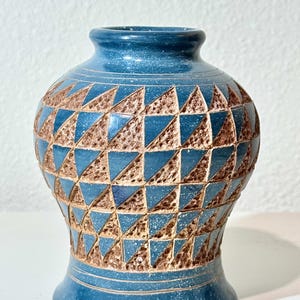 May include: A blue ceramic vase featuring a repeating geometric pattern of triangles. The design alternates between smooth, speckled blue triangles and textured, brown triangular indentations. The vase has a flared base and a narrow neck.