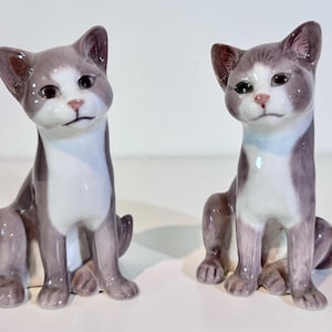 May include: Two ceramic cat figurines in a seated position. Each cat features a glossy finish with a grey and white color pattern, including a white chest, white muzzle, and grey ears, head, and back. The cats have dark eyes and pink noses.