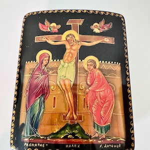May include: A rectangular black lacquer box featuring a hand-painted scene of the crucifixion with a central figure on a cross, flanked by two figures in robes. The artwork includes gold halos, two small winged figures, and Cyrillic text at the bottom.