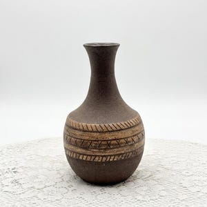 May include: A dark brown ceramic vase with a narrow neck and a rounded body. The midsection of the vase features two horizontal bands with etched, repeating diagonal and crisscross patterns in a lighter tan color, contrasting with the dark base.