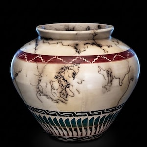 May include: A round ceramic vase with a cream-colored surface featuring black horsehair-style firing marks. The vase includes a horizontal band with a red zigzag pattern and a lower section decorated with a black Greek key design and teal feathers.