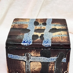 May include: A square ceramic box with a lid featuring a textured surface in shades of dark brown, black, and metallic gold. The design includes intersecting horizontal and vertical bands of mottled light blue glaze that create a grid-like pattern.