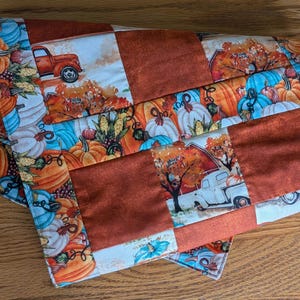 May include: A quilted table runner featuring a patchwork design of solid burnt orange fabric squares alternating with a printed fabric pattern of pumpkins, corn, and vintage trucks in shades of orange, blue, white, and brown on a light background.