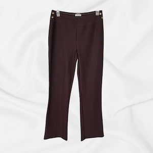 May include: Dark brown ribbed pants featuring a high-waisted design with two small gold-toned metal buttons on each side of the waistband. The pants have a straight-leg cut and a subtle vertical texture throughout the fabric.