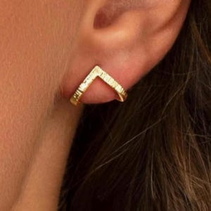 May include: A gold-toned ear cuff featuring a V-shaped design with a textured, hammered surface. The jewelry piece is positioned along the outer edge of the ear cartilage, showcasing a metallic finish and a distinct angular silhouette.