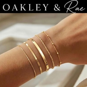 May include: A set of five gold-toned bracelets featuring varied designs including a thin box chain, a flat snake chain, a smooth polished bar, a twisted rope chain, and a delicate beaded chain, all displayed together on a wrist.