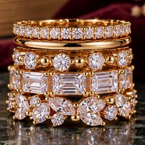 May include: A stack of five gold-toned rings featuring various clear gemstone cuts. The set includes a pavé band, a plain polished band, a round-cut stone band, an emerald-cut stone band, and a band with marquise-cut stones.