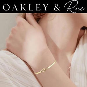 May include: A gold-toned bracelet featuring a smooth, thin band that terminates in a small, teardrop-shaped loop clasp. The text OAKLEY & Rae is displayed in white lettering against a black background at the top of the image frame.