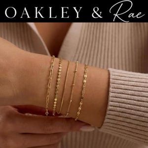 May include: A set of five gold-toned bracelets featuring distinct chain styles, including a paperclip link, a flat disc chain, a thin snake chain, a beaded chain, and a marquise-shaped link chain, displayed with the text OAKLEY & Rae above.