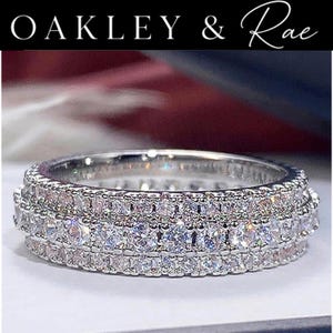 May include: A silver-toned band ring featuring three horizontal rows of clear, round-cut stones set in a metallic frame. The text OAKLEY & Rae is displayed in white lettering at the top of the image against a dark background.