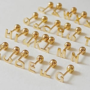 May include: A collection of gold-toned metal stud earrings, each featuring a single uppercase letter of the alphabet. Every earring consists of a flat letter charm attached to a thin post with a small, spherical ball backing.