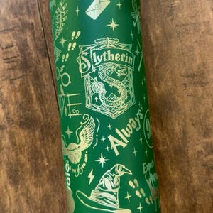 Puede incluir: Una botella de agua cilíndrica de color verde con un estampado gráfico en amarillo claro del escudo de la casa Slytherin, un sombrero seleccionador, una bola alada, un sobre, estrellas y el texto Always. Incluye varios iconos mágicos.