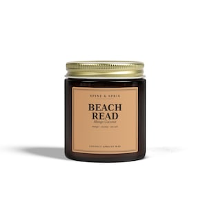 Peut inclure: Une bougie en pot de verre ambré foncé avec un couvercle en métal doré. L'étiquette rectangulaire porte le texte SPINE & SPRIG, BEACH READ, Mango Coconut, mango coconut sea salt et COCONUT APRICOT WAX imprimé en noir sur fond beige.