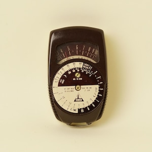 May include: A dark brown vintage Tower light meter featuring a circular dial with white markings, numbers, and text including "LIGHT VALUE," "ADJ. TO ZERO," and "SET LIGHT VALUE TO ASA INDEX." The device has a curved, rectangular plastic casing.