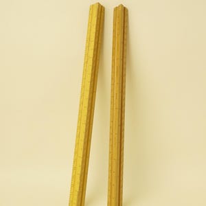 May include: Two yellow triangular architectural scales standing upright. Each scale features three distinct sides with printed black measurement markings in inches and fractional increments, designed for precise technical drafting and engineering tasks.