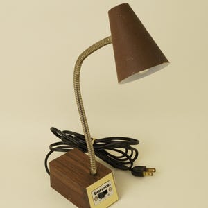 May include: A brown desk lamp with a conical shade, a flexible metallic gooseneck arm, and a rectangular base with a wood-grain finish. The base features a white control panel with the brand name tensor and a black power switch with settings.