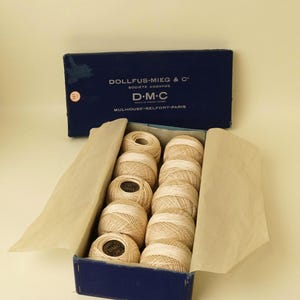 May include: A dark blue rectangular box labeled DOLLFUS-MIEG & C D.M.C MULHOUSE-BELFORT-PARIS containing ten round balls of beige cotton thread arranged in two rows. The box is lined with light tan tissue paper that folds over the sides.