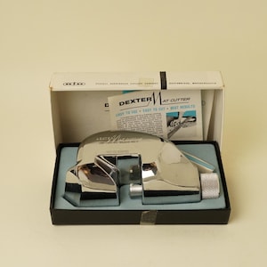 May include: A chrome-plated Dexter Mat Cutter tool housed in a black box with a light blue foam insert. The packaging includes a white instruction card with the text Dexter Mat Cutter, Easy to Use, Easy to Cut, Best Results, and Russell Harrington Cutlery Company.